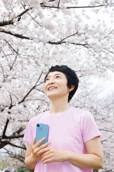 亚洲女人带着智能手机停在有樱花的公园里 — 图库照片