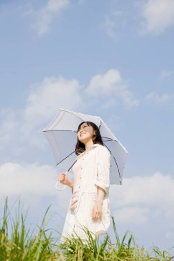 Beyaz şemsiyeli mutlu genç bir kadın mavi gökyüzüne karşı çimenli çayırlarda yürüyor.