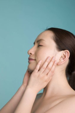 Genç Asyalı gözleri kapalı bir kadın mavi arka planda tek başına yüzüne dokunuyor. 