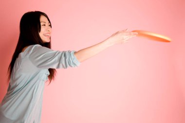 Mutlu genç Asyalı kadın pembe arka planda frizbi atıyor.