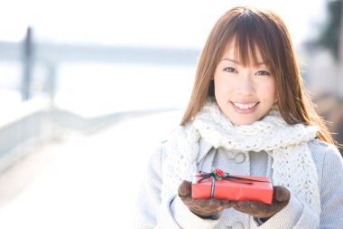 Kışlık giysiler içinde güzel bir Asyalı kadın hediye kutusu tutuyor ve gülümsüyor. 