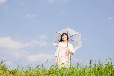 Güneşli bir günde yeşil çayırda yürüyen şemsiyeli genç bir kadın.