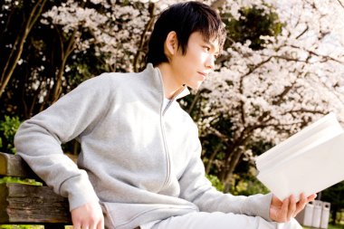 Genç Asyalı adam bahar parkında kitap okuyor. 