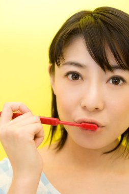 Asyalı genç bir kadın sarı arka planda dişlerini fırçalıyor.