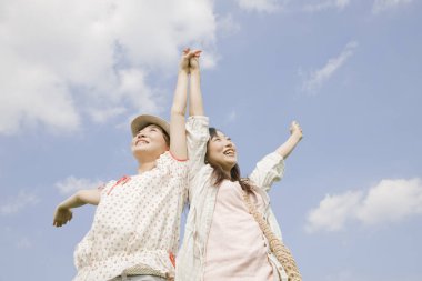 Kaygısız Asyalı genç kadınlar el ele tutuşup mavi gökyüzüne karşı zıplıyorlar.