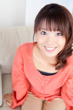 Güzel genç Asyalı kadın evde oturmuş kameraya gülümsüyor.