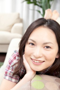 Genç Asyalı kadın koltukta yastığa uzanmış kameraya gülümsüyor. 