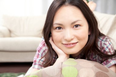 Genç Asyalı kadın koltukta yastığa uzanmış kameraya gülümsüyor. 