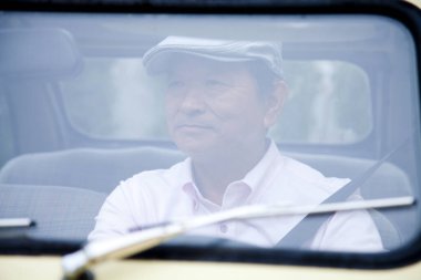 Şapkalı yaşlı Asyalı adam arabayı yakından sürüyor. 