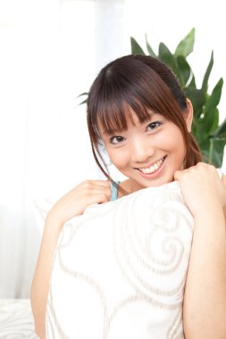 Güzel genç Asyalı kadın yastığa sarılıyor ve evde kameraya gülümsüyor.