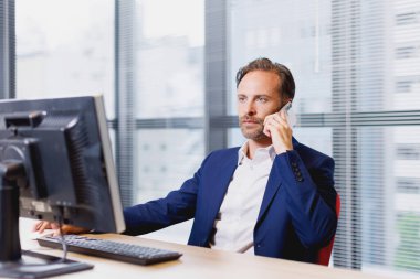 Modern ofiste bilgisayar üzerinde çalışan mavi takım elbiseli yakışıklı genç iş adamı.