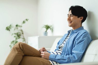 Evdeki kanepede oturan, günlük kıyafetler giyen Asyalı genç adam.