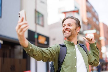 Yeşil tişörtlü genç beyaz adam cep telefonuyla selfie çekiyor.