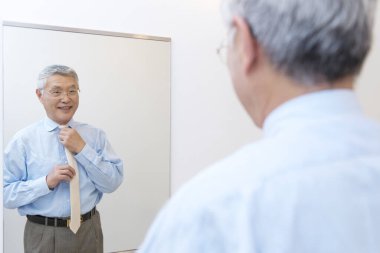 Asyalı kıdemli adam aynaya bakıyor ve gülümsüyor, aynanın önünde duruyor.