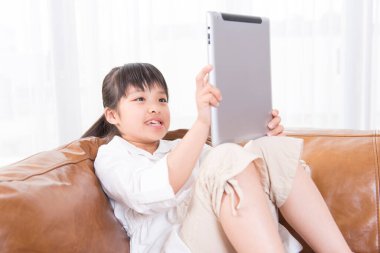 Küçük kız beyaz koltukta tablet kullanıyor.