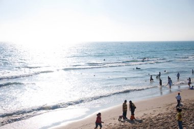 İnsanların okyanustan ve parlak gökyüzünün altında dalgalardan keyif aldığı bir kumsal sahnesi.