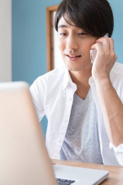 Genç adam dizüstü bilgisayar kullanıyor ve akıllı telefondan konuşuyor. 