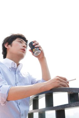 Genç Asyalı adam dışarıda sigara içiyor ve içiyor. 