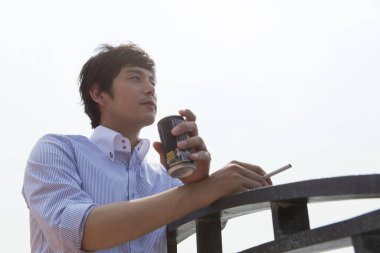 Genç Asyalı adam dışarıda sigara içiyor ve içiyor. 