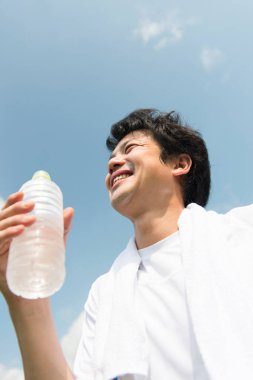 Genç Asyalı adam parkta antrenmandan sonra şişeden içiyor.