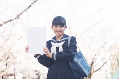 Gülümseyen Japon öğrenci elinde boş beyaz posterle caddede çiçek açan kiraz ağaçlarıyla