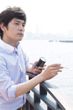 Genç Asyalı adam dışarıda sigara içiyor ve içiyor. 