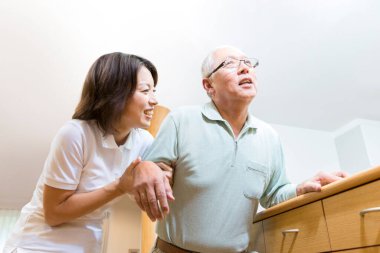 Asyalı kadın bakıcı yaşlı adamın yürümesine yardım ediyor.