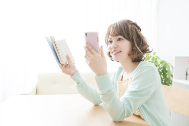 Kitaplı genç Asyalı kadın evde akıllı telefon kullanıyor. 