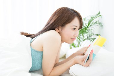 Genç Asyalı kadın evde beyaz bir yatakta kitap okuyor.