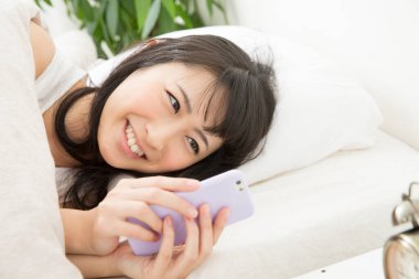 Asyalı genç kadın yatakta akıllı telefon kullanıyor. 
