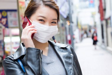 Maske takan Asyalı kadın sokakta akıllı telefondan konuşuyor.