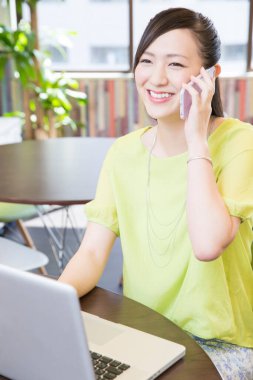 Asyalı iş kadını laptopta çalışıyor ve ofisteki akıllı telefondan konuşuyor.