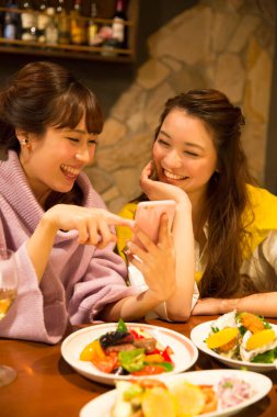 İki genç Asyalı kadın restoranda yemek yiyor, telefonda bir şeyler izliyorlar.