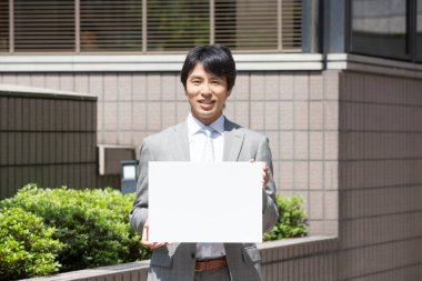 Asyalı yakışıklı, profesyonel bir iş adamı elinde boş bir levhayla sokakta kameraya gülümsüyor. 