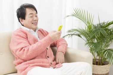 Asyalı kıdemli bir adam evdeki koltukta otururken akıllı telefon kullanıyor.