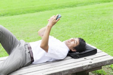 gülümseyen Asyalı iş adamı parkta bankta uzanıyor ve akıllı telefon kullanıyor.