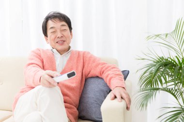 Asyalı kıdemli adam koltukta oturuyor ve uzaktan kumanda kullanıyor. 