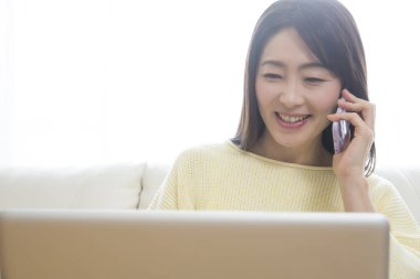 Genç Asyalı kadın dizüstü bilgisayarda çalışıyor ve evde akıllı telefonla konuşuyor. 