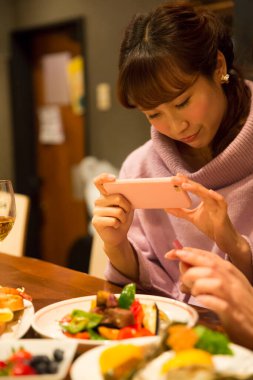 İki genç Asyalı kadın restoranda yemek yiyor, telefonda bir şeyler izliyorlar.