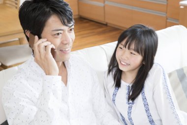 Kanepede kızıyla otururken akıllı telefondan konuşan Asyalı bir adam. 