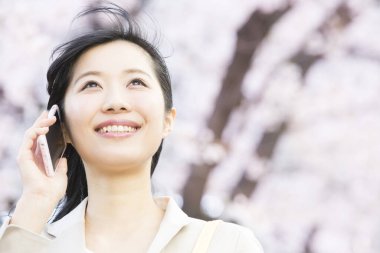 Güzel genç Asyalı kadın akıllı telefonla konuşuyor ve bahar parkına bakıyor.  