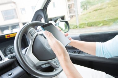 Genç bir kadın araba kullanıyor ve elinde akıllı telefon tutuyor. 