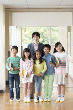 Okul üniformalı bir grup öğrenci okul koridorunda öğretmenle birlikte duruyorlar.