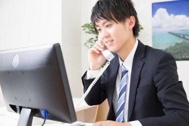 Genç Asyalı işadamı ofiste çalışırken telefon ve bilgisayar kullanıyor.