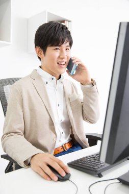 Genç adam ofiste bilgisayarla çalışıyor ve akıllı telefondan konuşuyor. 