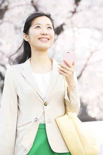 漂亮的笑着的年轻亚洲女人 拿着手提包 用智能手机走在春天的公园里 — 图库照片
