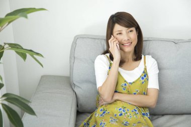 Asyalı genç bir kadın evdeki koltukta cep telefonu kullanıyor.