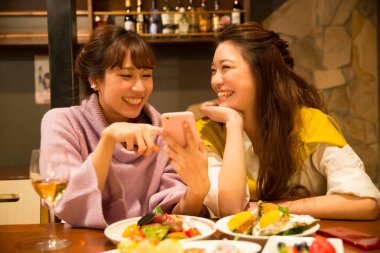 İki genç Asyalı kadın restoranda yemek yiyor, telefonda bir şeyler izliyorlar.