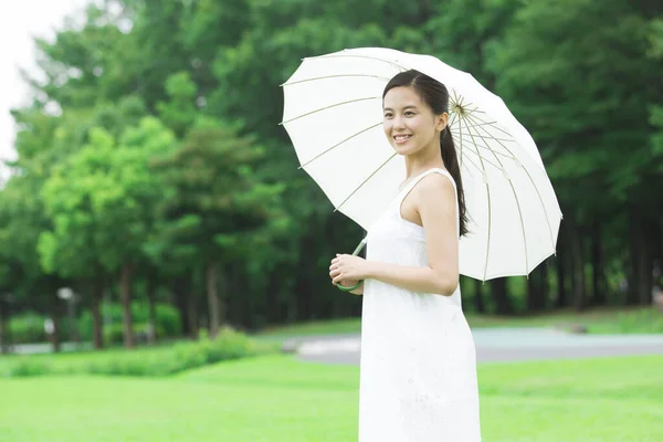 年轻的日本女人带着白色的雨伞在绿色的夏季公园里散步 身着白衣的年轻女子的白天肖像 — 图库照片