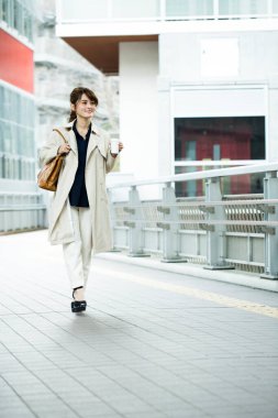 Genç Asyalı kadın elinde kahveyle kaldırımda yürüyor.